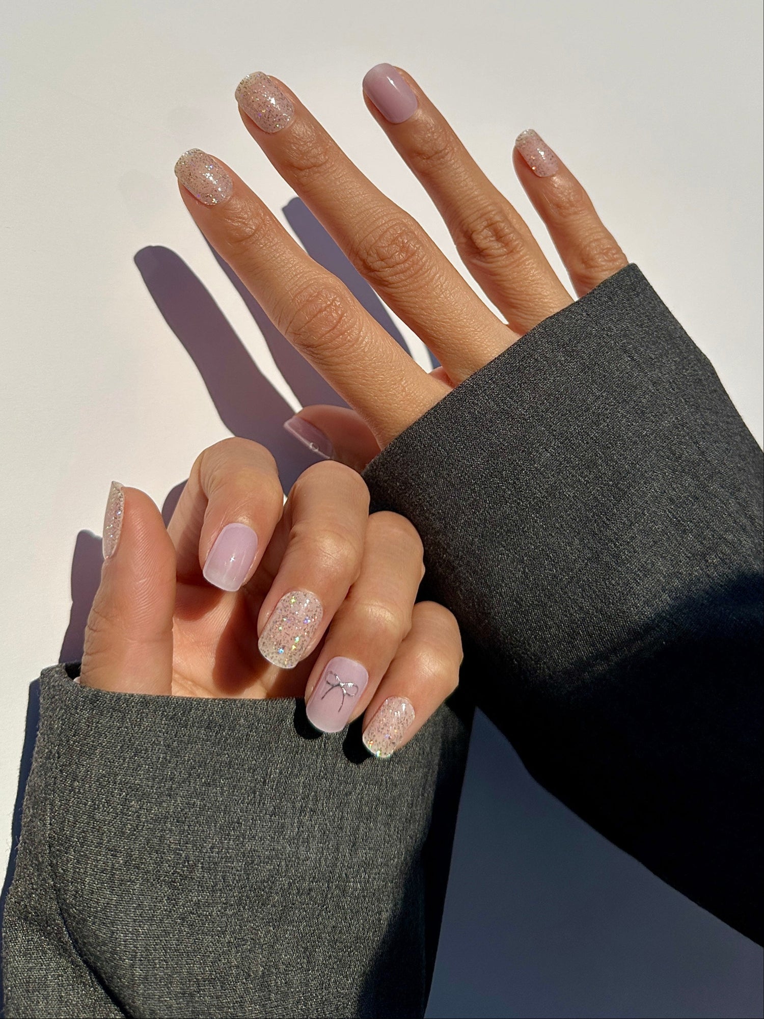 Hand modeling of semicured gel stickers in varying shades of lilac and purple with shimmery silver glitter and silver foil bow details.