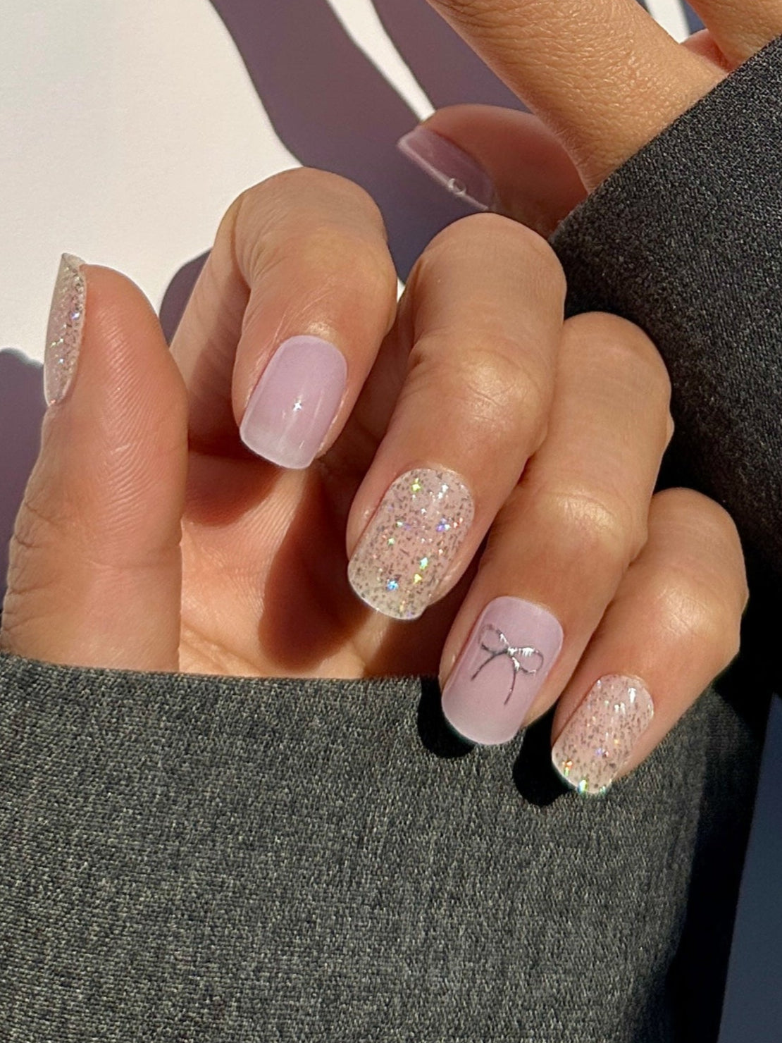 Hand model of semicured gel stickers in varying shades of lilac and purple with shimmery silver glitter and silver foil bow details.