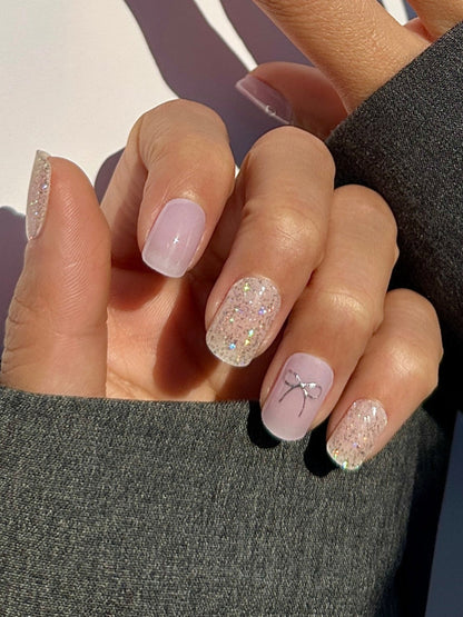 Hand model of semicured gel stickers in varying shades of lilac and purple with shimmery silver glitter and silver foil bow details.